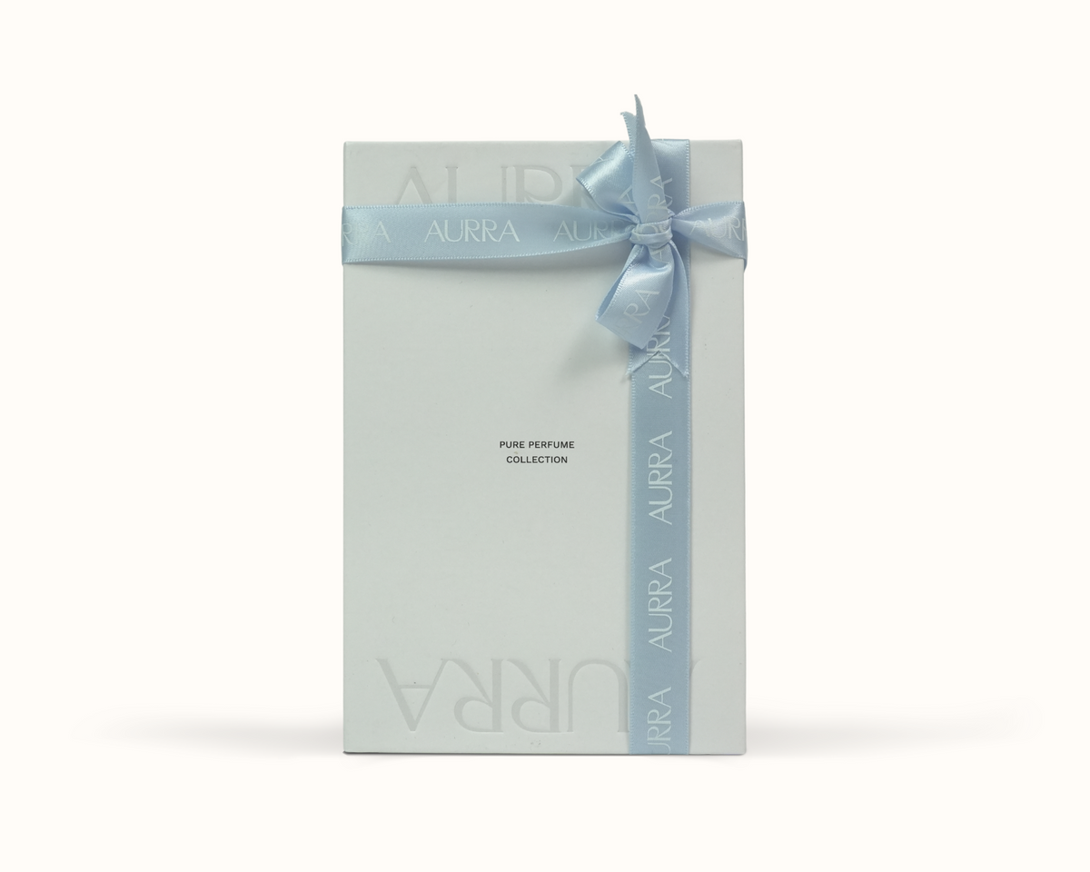 A tall, rectangular white perfume discovery set from AURRA, tied with a light blue satin ribbon and bow, set against a plain off-white background.