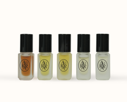 Five small cylindrical bottles with black caps and frosted glass, arranged in a row on a light gray background. Each bottle contains a different colored liquid: orange, light brown, pale yellow, clear, and milky white.